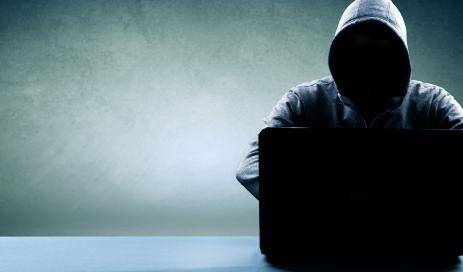 Eine Cyberattacke durchführender Hacker Angreifender Hacker, der im Dunkeln mit übergezogenem Kapuzenpullover vor einem Notebook sitzt und versucht, Passwörter einer Nutzerdatenbank zu knacken.