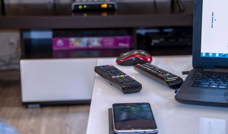 Engity's DeviceAuth (Keyboardless Device Login) A TV screen with a set-top box in the background which are steered by two TV remotes and a smartphone lying on a table; a login to a content service is performed by the user.