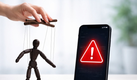 Eine Marionette aus dunklem Holz, die für Social Engineering steht und von einer Hand bewegt wird, daneben ein Smartphone mit einem Warnhinweis auf dem Display, welches den Angriff auf ein 2-Faktor-Zugangssystem symbolisiert.